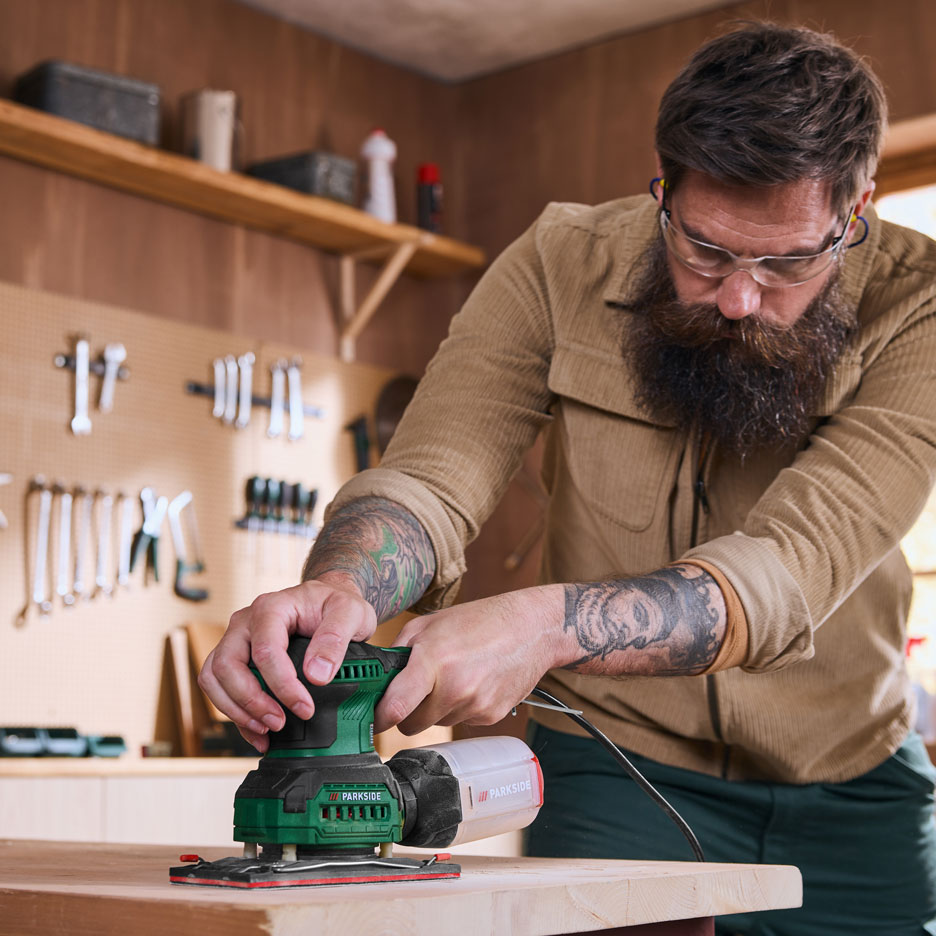 Un homme dans un atelier ponce une planche de bois avec une ponceuse électrique.
