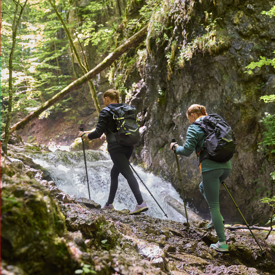 Twee vrouwen met rugzakken en trekkingstokken wandelen langs een waterval.