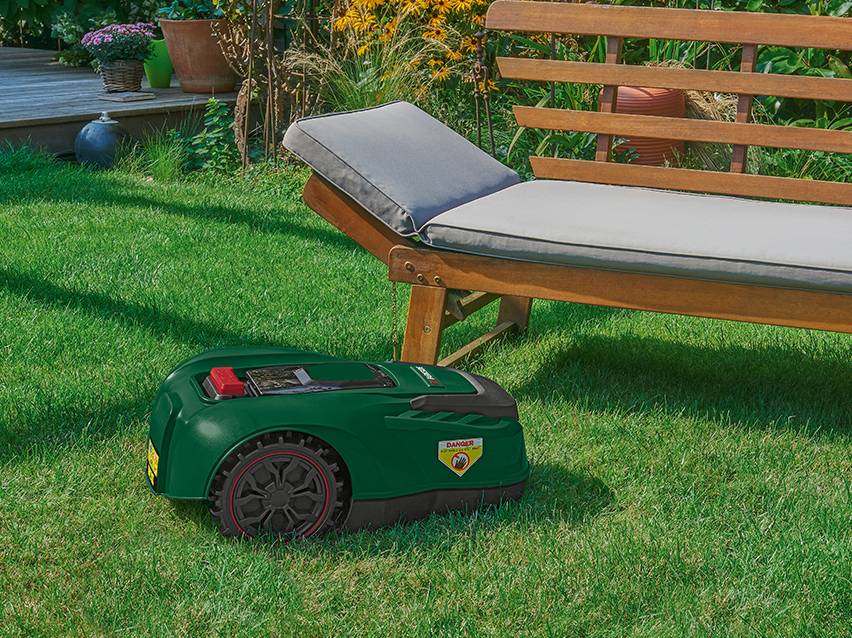 Tondeuse robot verte sur une pelouse, avec une chaise longue en bois en arrière-plan.