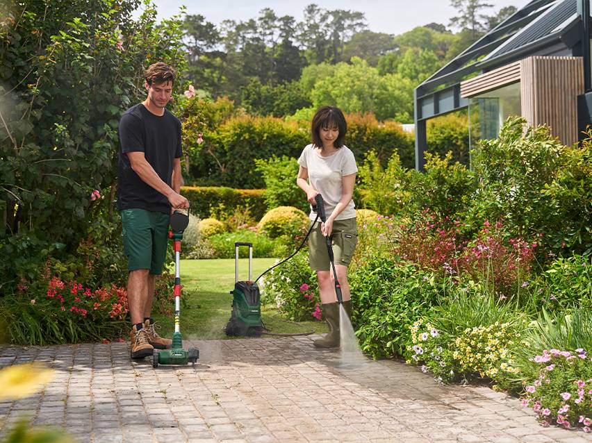 Man en vrouw reinigen een oprit met een terrasreiniger en hogedrukreiniger in een tuin.