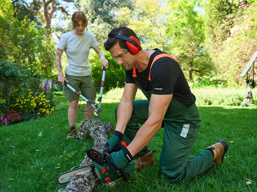 Man zaagt boomstam met kettingzaag, vrouw snoeit takken met takkenschaar in de tuin.