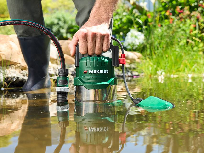 Persoon met laarzen in water, houdt een groene dompelpomp vast met een aangesloten slang.