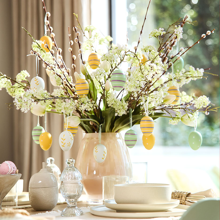 Vaas met wilgentakken, witte bloemen en kleurrijke paaseieren op een gedekte tafel.