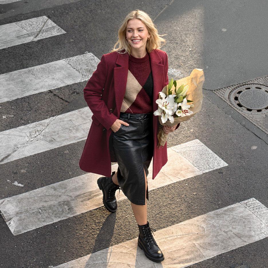 Lachende vrouw in bordeauxrode jas, argyle trui en leren rok, steekt de straat over met een boeket lelies.