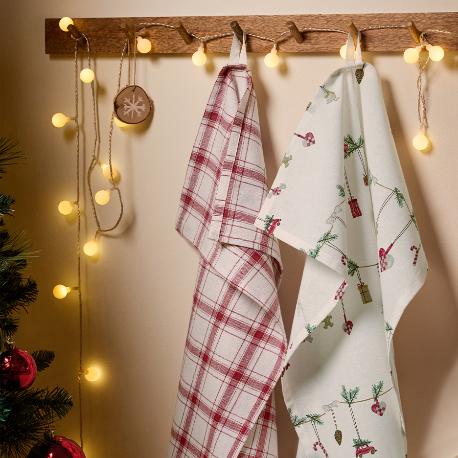 Twee feestelijke kerstkeukenhanddoeken, één geruit en één met kerstmotieven, hangend aan een houten kapstok met lichtsnoer.