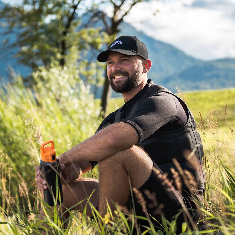 Lachende man met zwarte pet en neopreen vest, houdt een oranje waterfles vast.