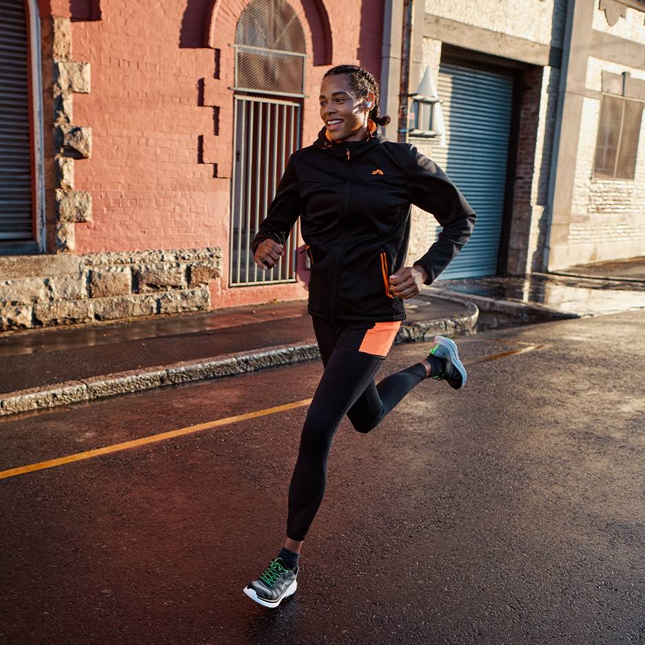 Man in zwart hardloopjack en legging met oranje accenten, rennend op een natte straat.
