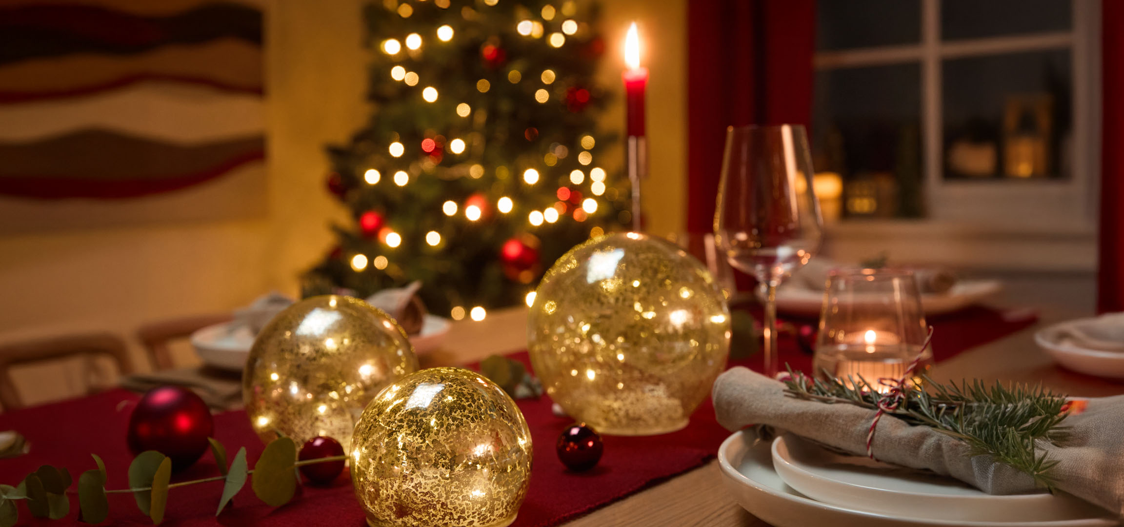 Kersttafeldecoratie met gouden lichtbollen, rode kerstballen en dennentakjes op een rood tafelkleed.
