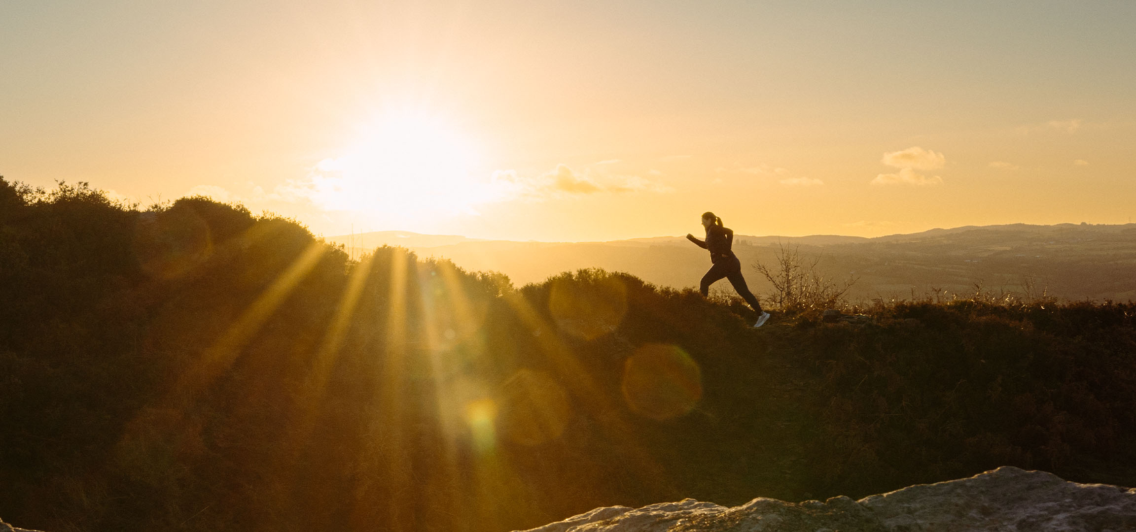 Femme courant sur une colline au coucher du soleil, avec des rayons de soleil et un paysage.