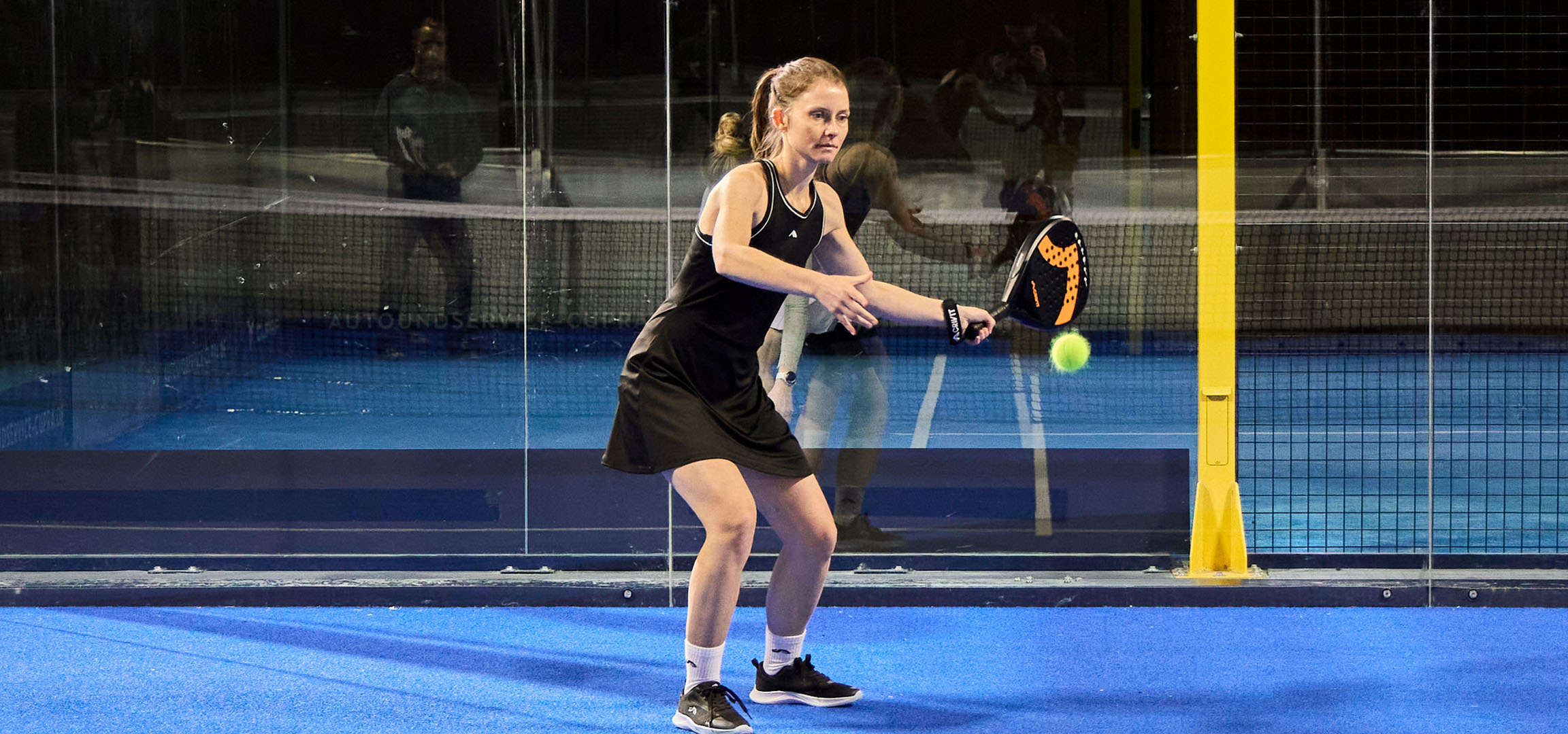Vrouw speelt padel op een blauwe baan, gekleed in zwarte sportkleding.