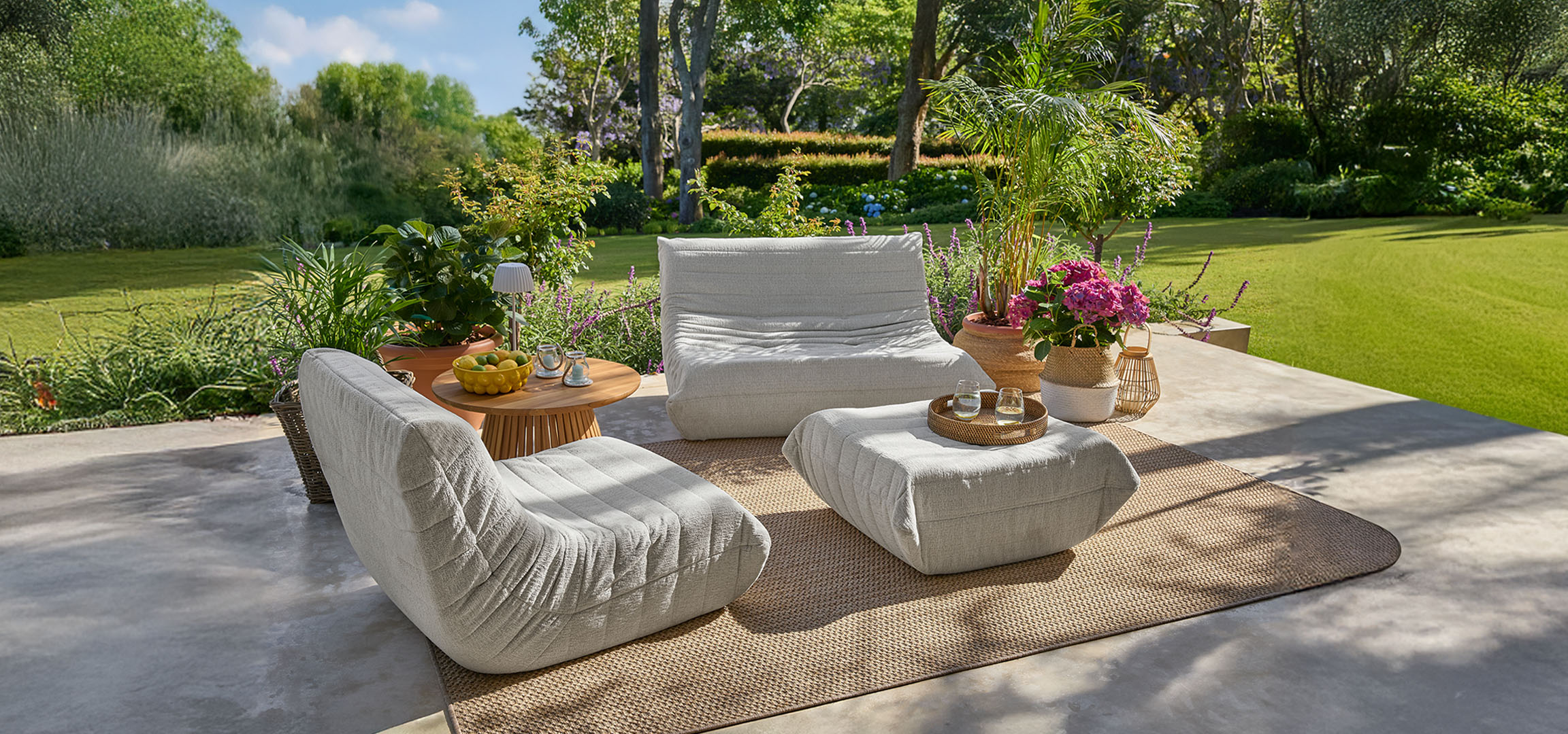 Lichtgrijze fauteuils en poefs op een terras met een houten tafel en groen gazon.