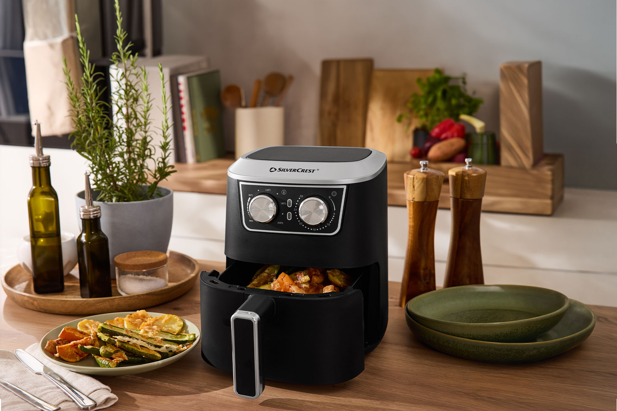 Zwarte airfryer met groenten, olie en kruiden op een houten tafel.
