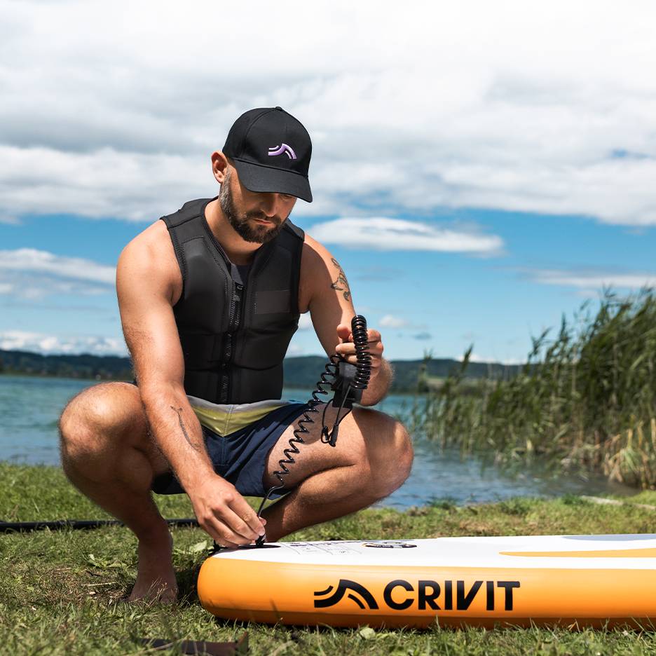Man bevestigt veiligheidslijn aan CRIVIT SUP-board, draagt zwemvest en pet.