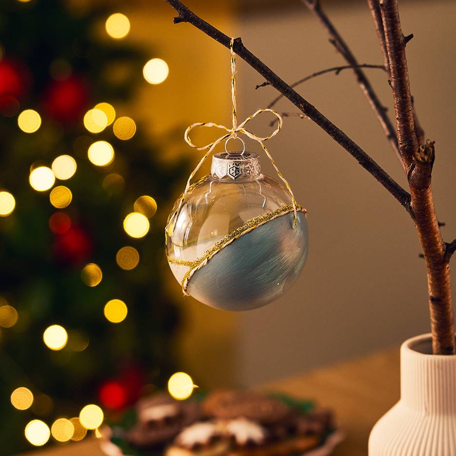 Transparante en blauwe kerstbal met gouden glitter, hangend aan een tak met kerstverlichting op de achtergrond.