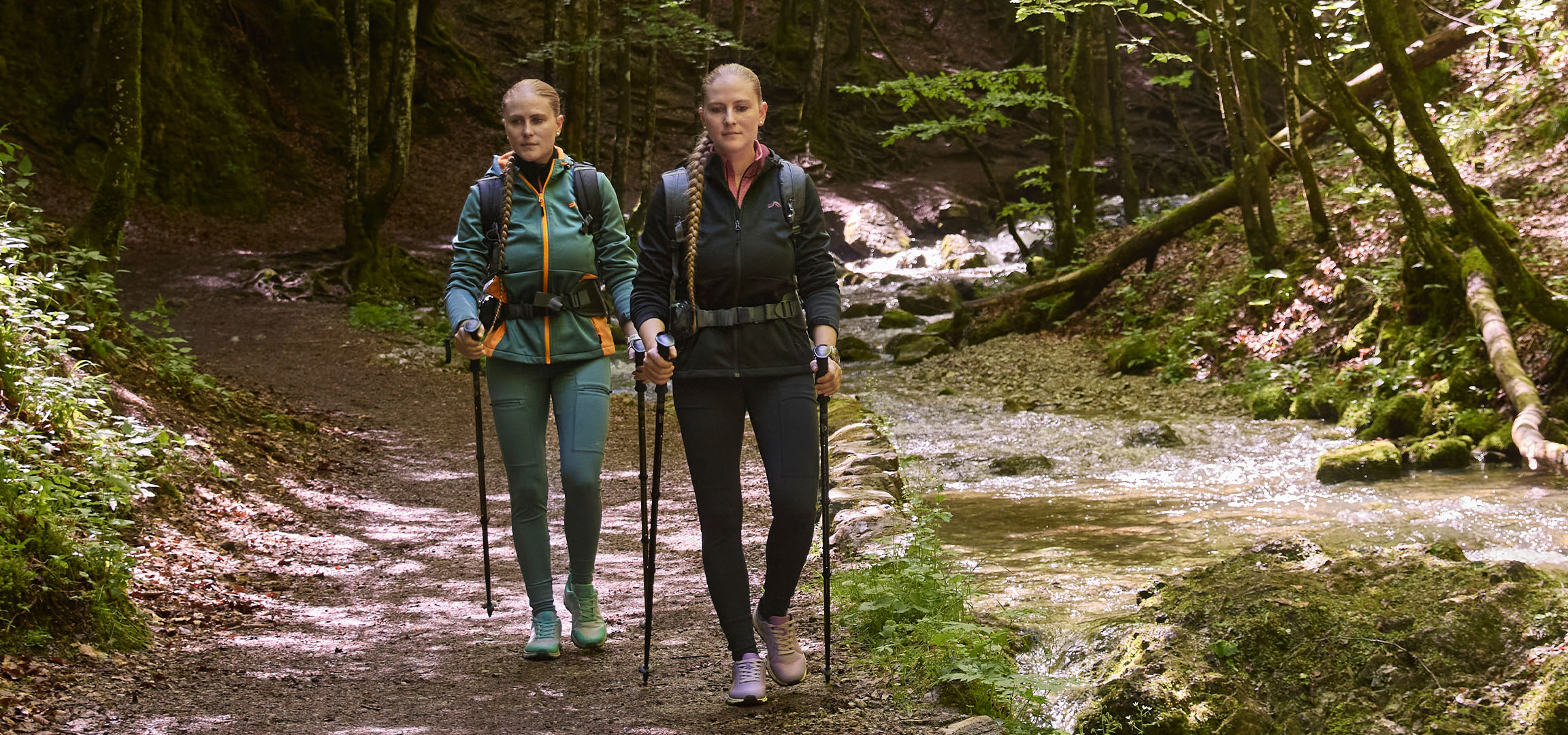 Deux femmes font de la randonnée le long d'un ruisseau dans une forêt avec des bâtons de marche.