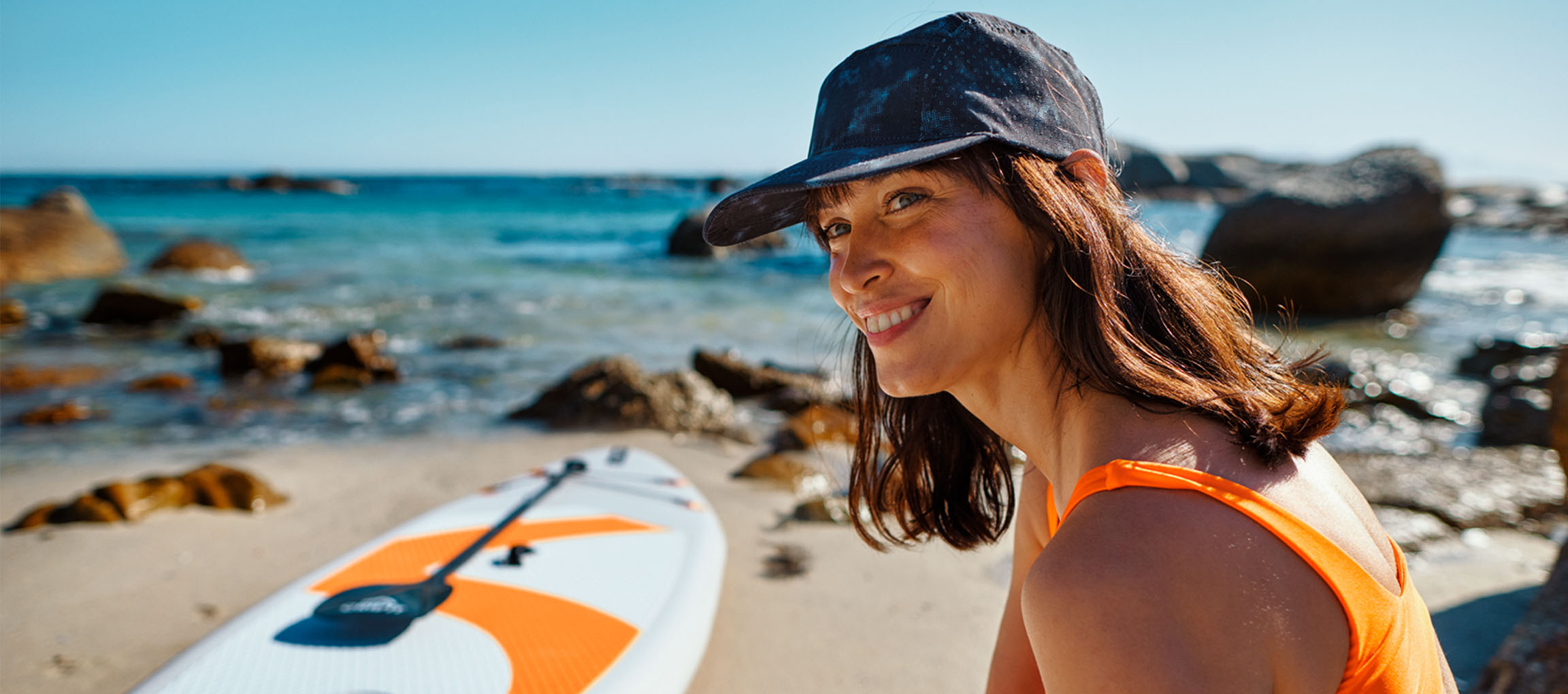 Lachende vrouw in een oranje badpak en pet, naast een paddleboard op het strand.