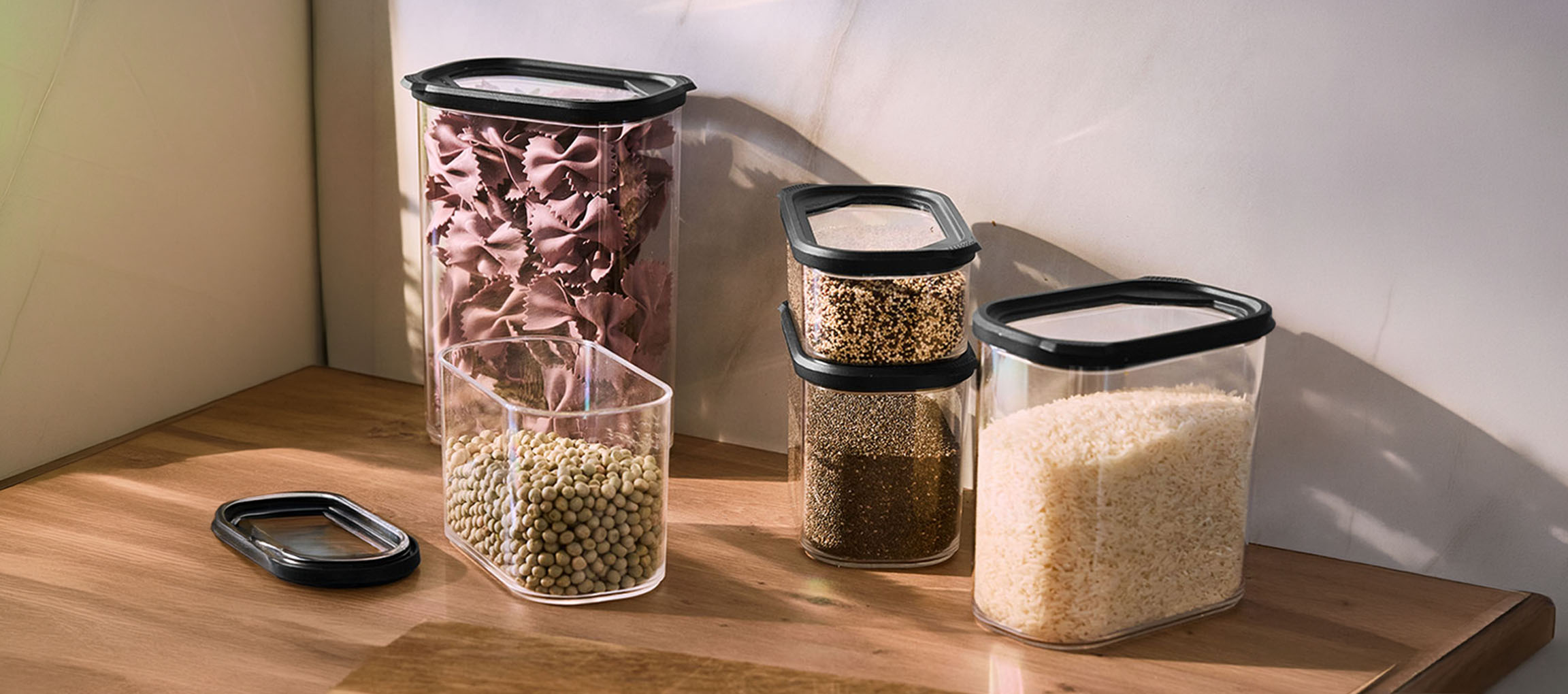 Transparante keukencontainers met zwarte deksels, gevuld met pasta, linzen, quinoa, chiazaad en rijst op een houten oppervlak.