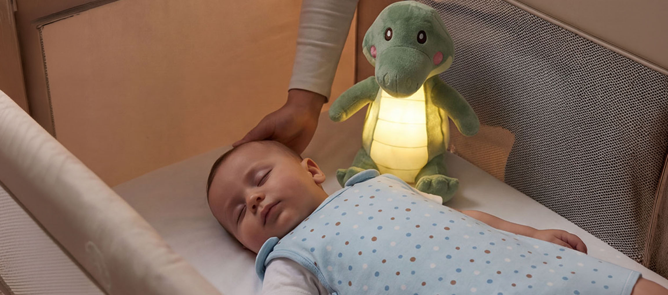 Bébé dormant dans un berceau avec une veilleuse en peluche, un adulte caressant sa tête.