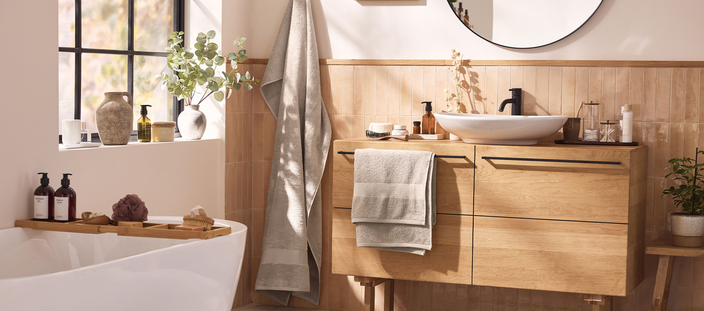 Une salle de bain moderne avec un meuble-lavabo en bois, un lavabo blanc, un miroir rond et une baignoire avec accessoires.