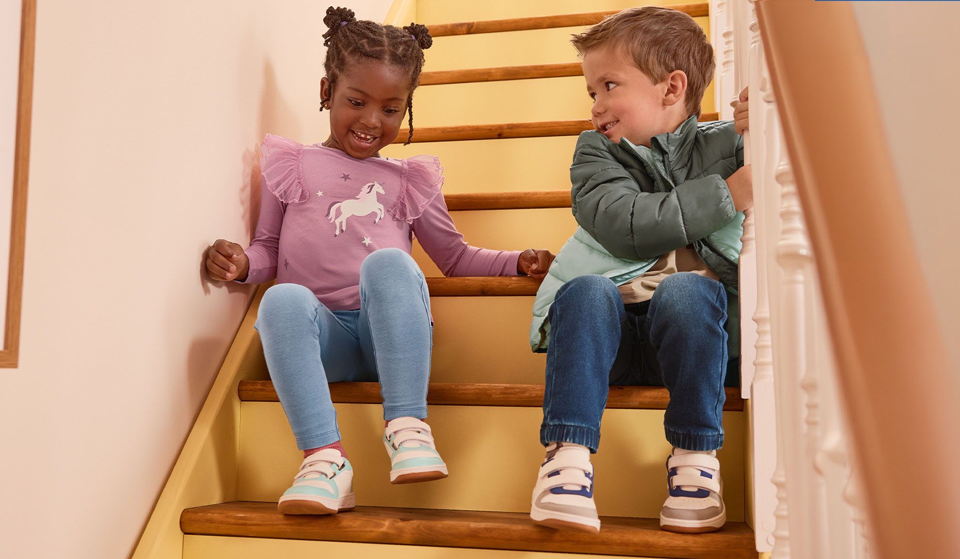 Twee kinderen in casual kleding en sneakers zitten op een houten trap, lachend naar elkaar.