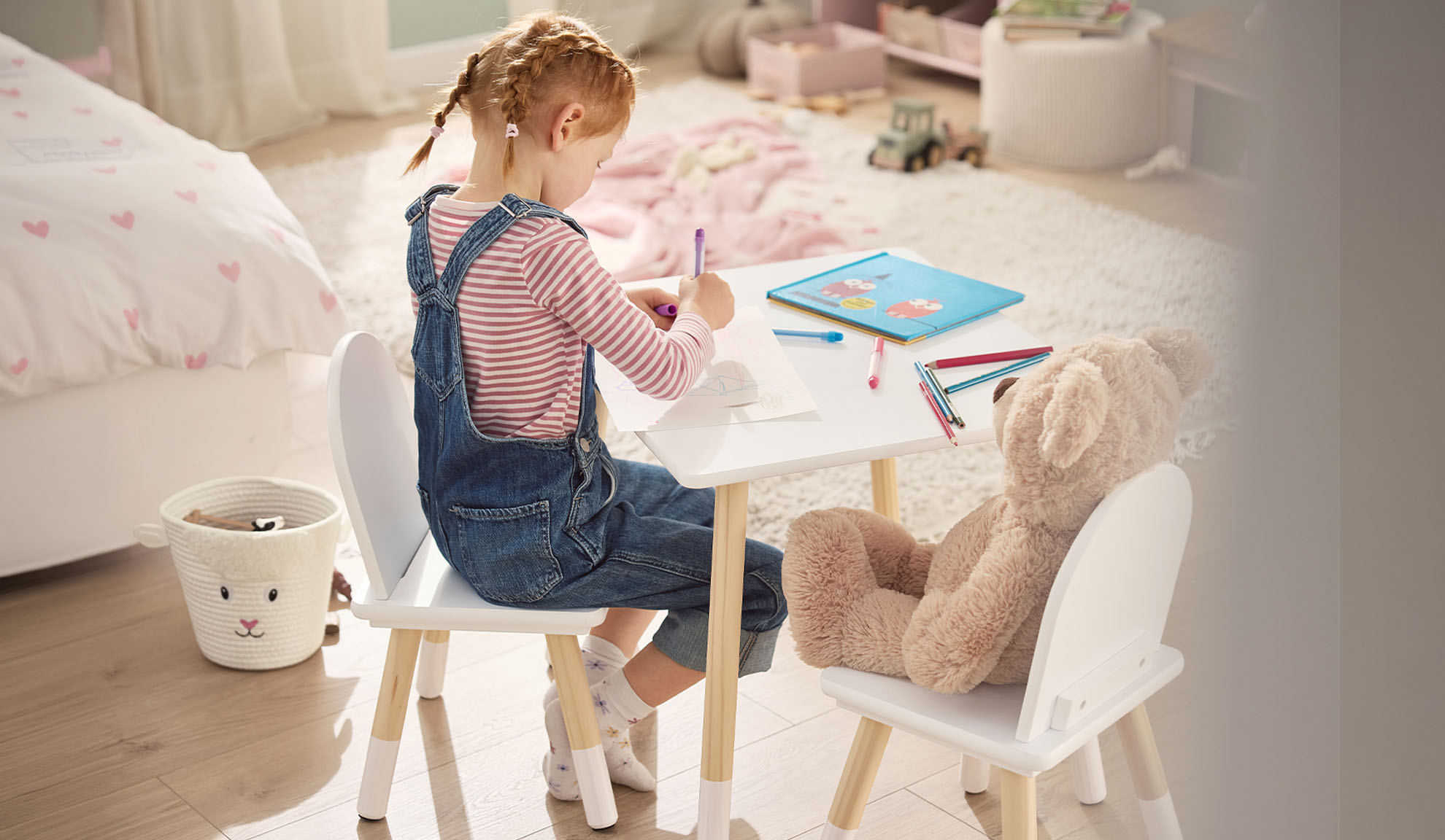 Fille dessinant à une table blanche avec un ours en peluche, dans une chambre d'enfant.