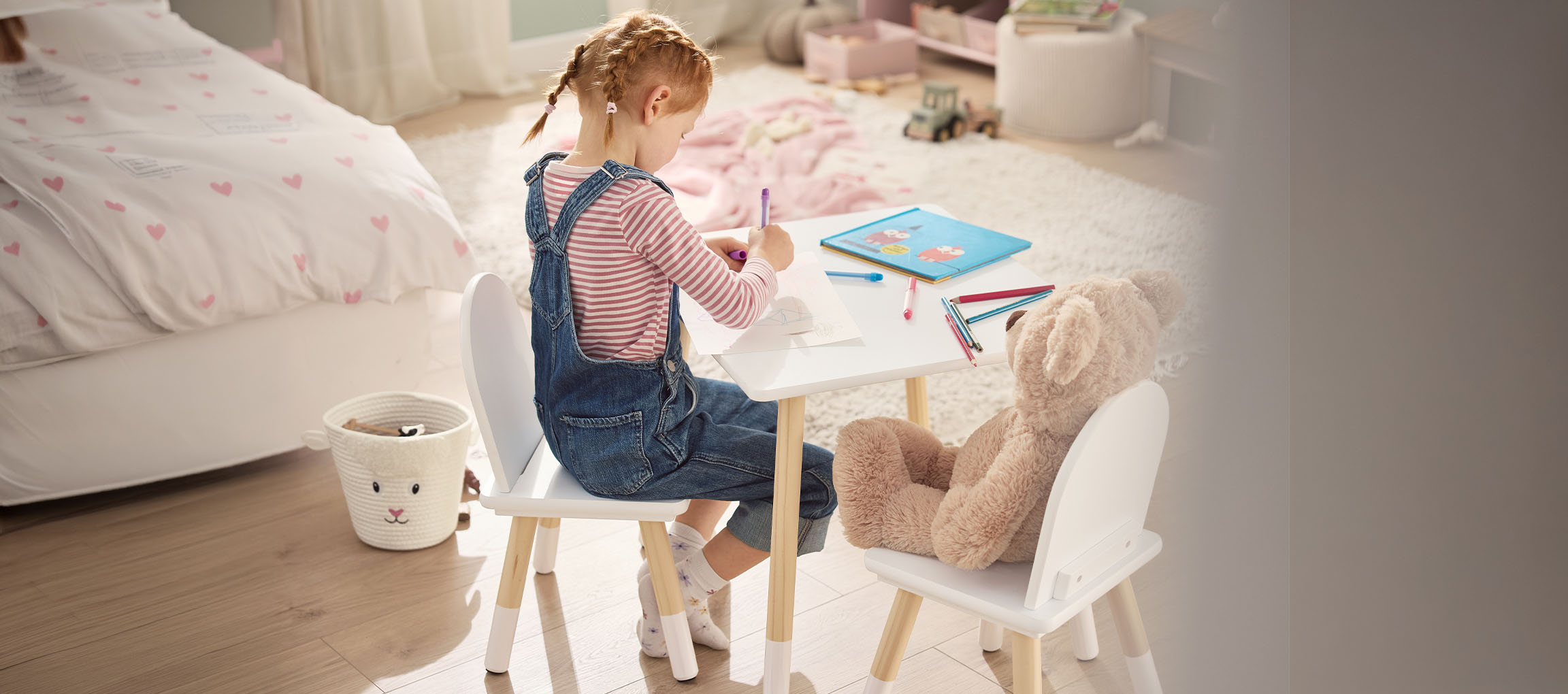 Fille dessinant à une table blanche avec des chaises, un ours en peluche à côté d'elle.