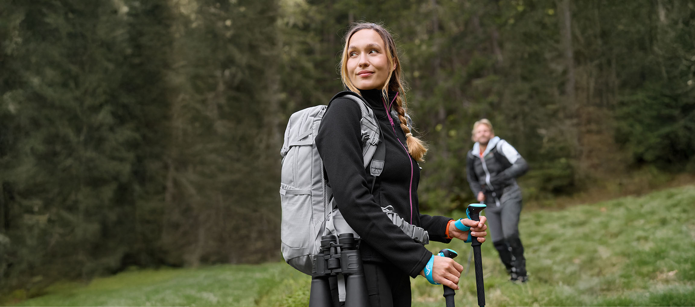 Femme avec sac à dos et bâtons de randonnée, avec un homme en arrière-plan, dans une forêt.