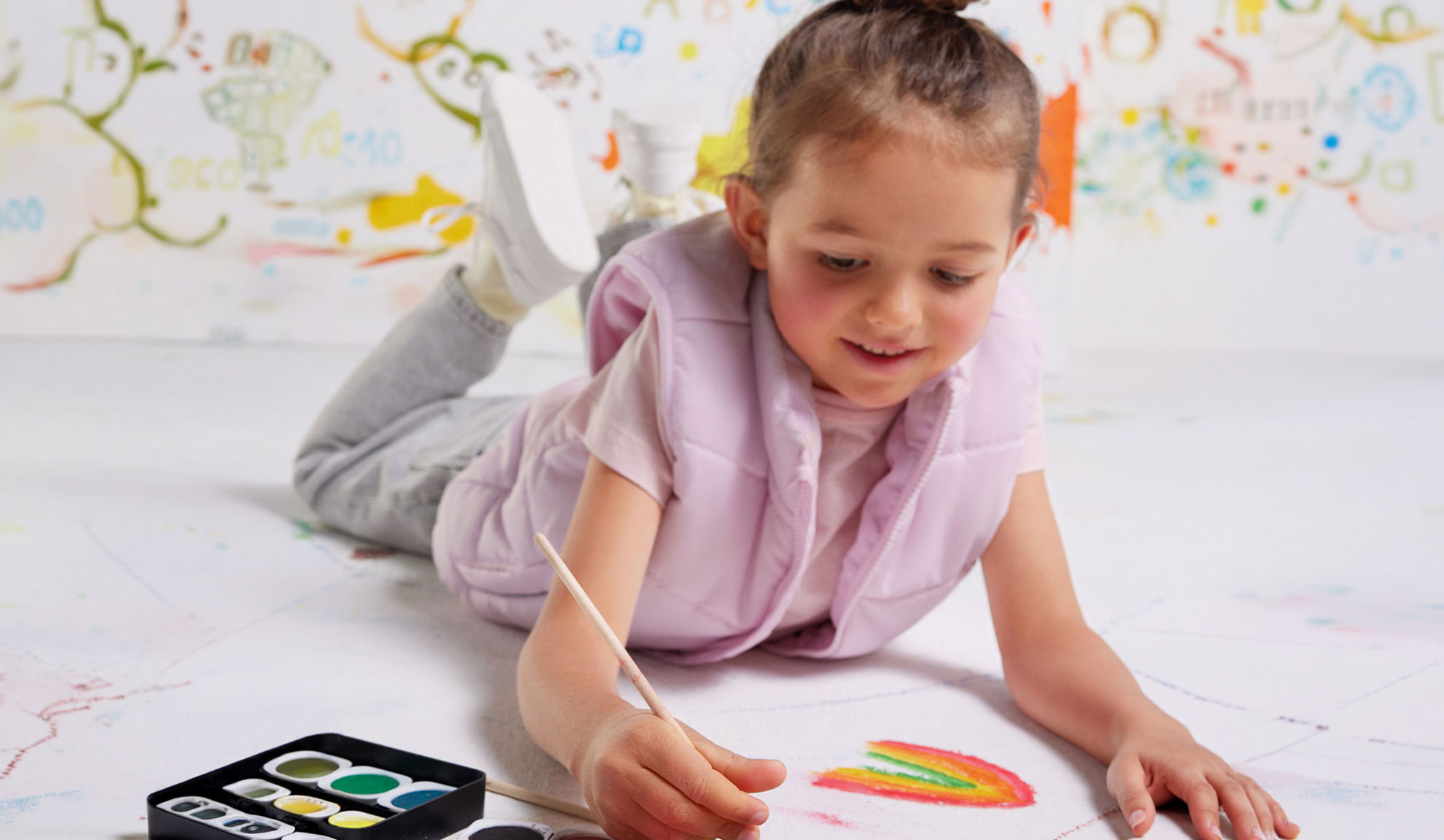 Une enfant peint un arc-en-ciel sur papier avec des aquarelles, vêtue d'un gilet rose.