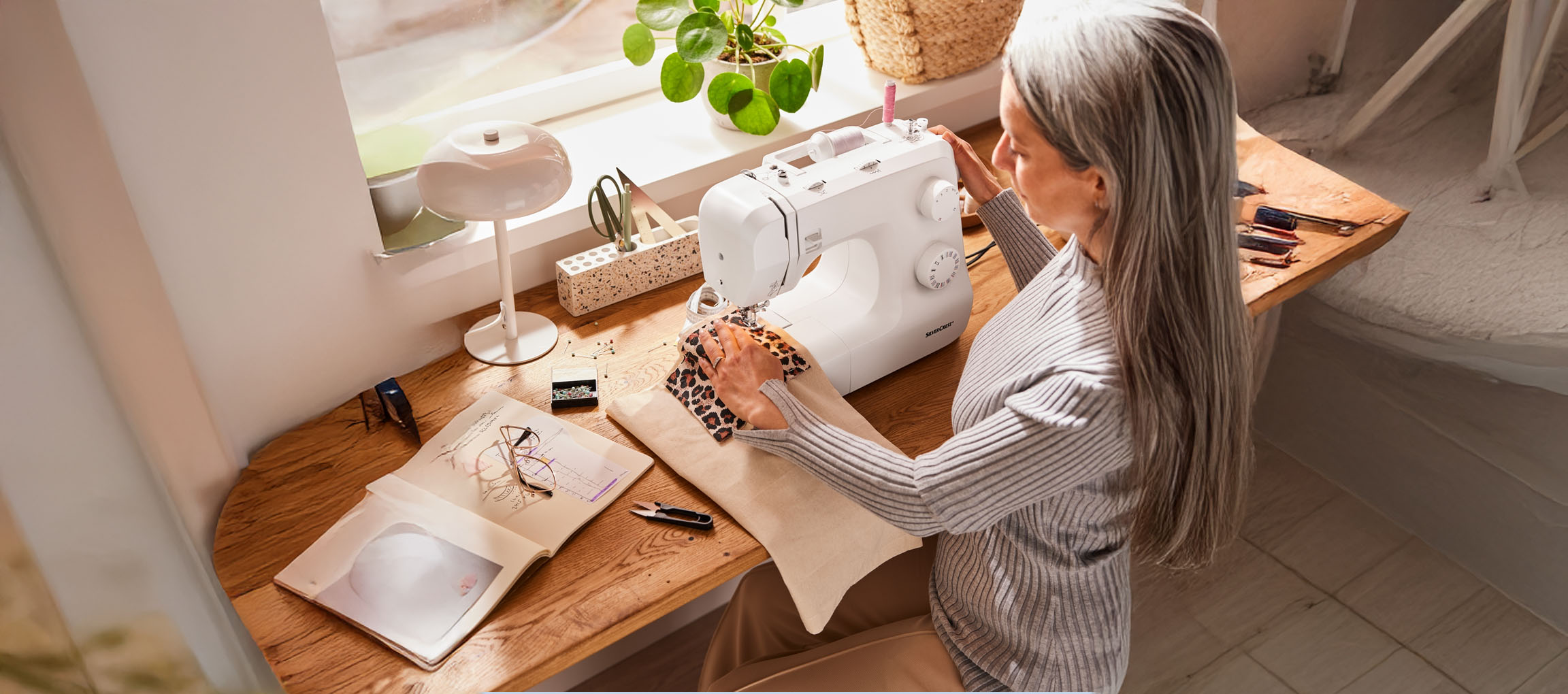 Une femme coud sur une machine à coudre SilverCrest, avec un carnet de croquis et des accessoires de couture sur la table.
