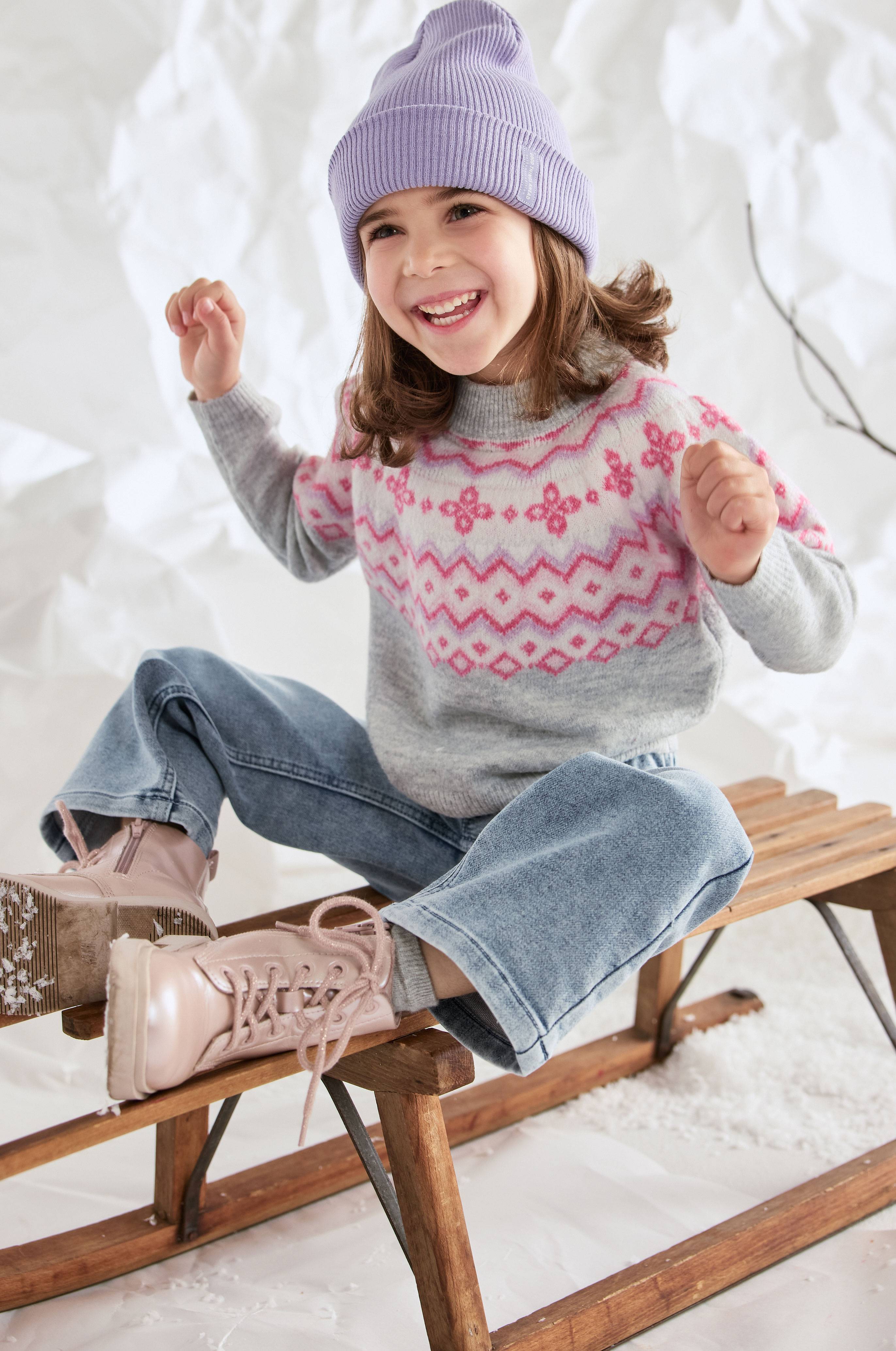 Meisje met paarse muts, gebreide trui met Noors patroon en wijde jeans, zittend op een houten slee.