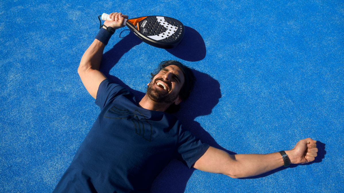 Homme souriant allongé sur un court de padel bleu, raquette de padel au-dessus de la tête.