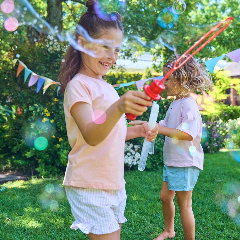 Twee meisjes spelen met bellenblaas in de tuin, met vrolijke vlaggetjes op de achtergrond.
