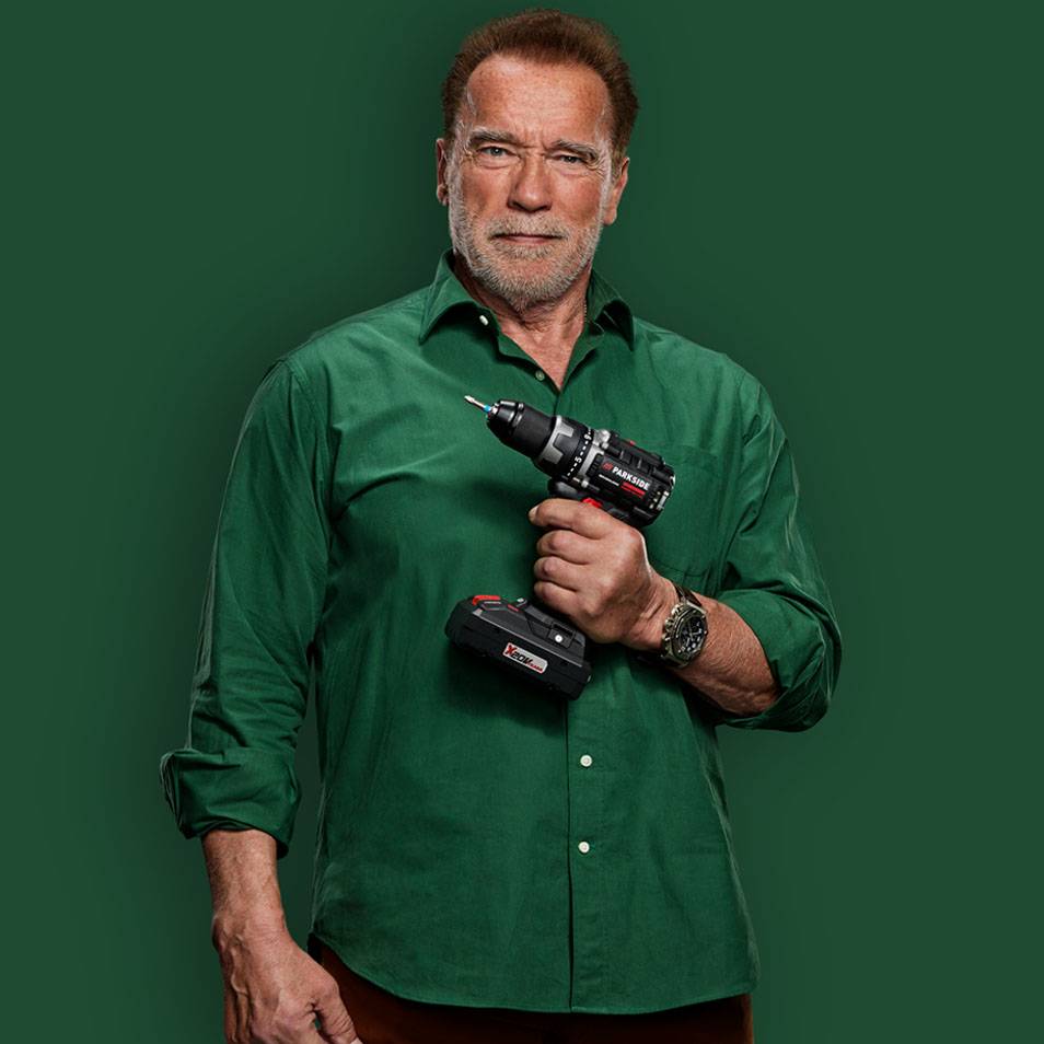 Arnold Schwarzenegger in een groen overhemd met een boormachine in zijn hand.