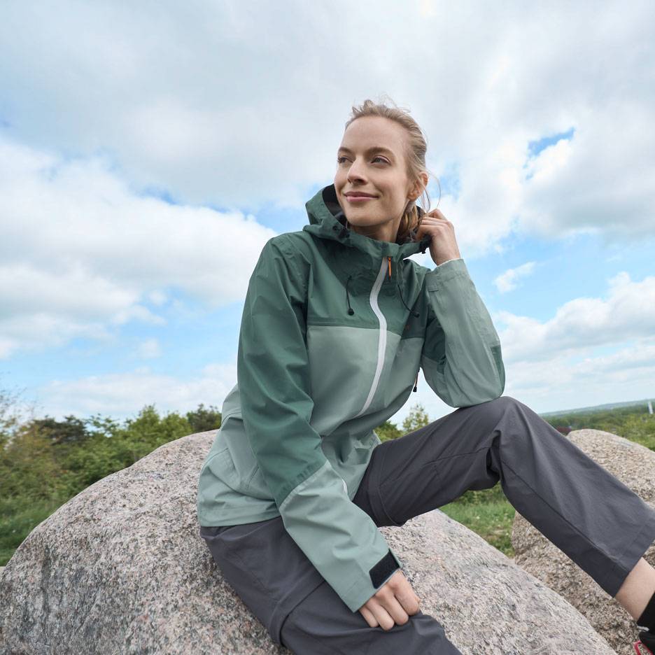 Vrouw in een groene outdoorjas en grijze broek zit op een rots, kijkend in de verte.