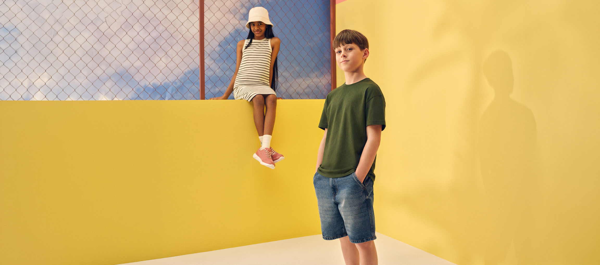 Twee kinderen in casual zomerkleding: een meisje in een gestreepte jurk en een jongen in een groen T-shirt en spijkershort.