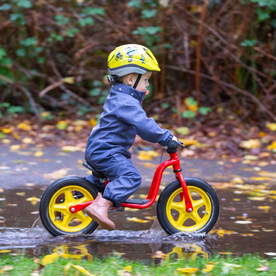 Kind op loopfiets in een plas, met helm en overall.