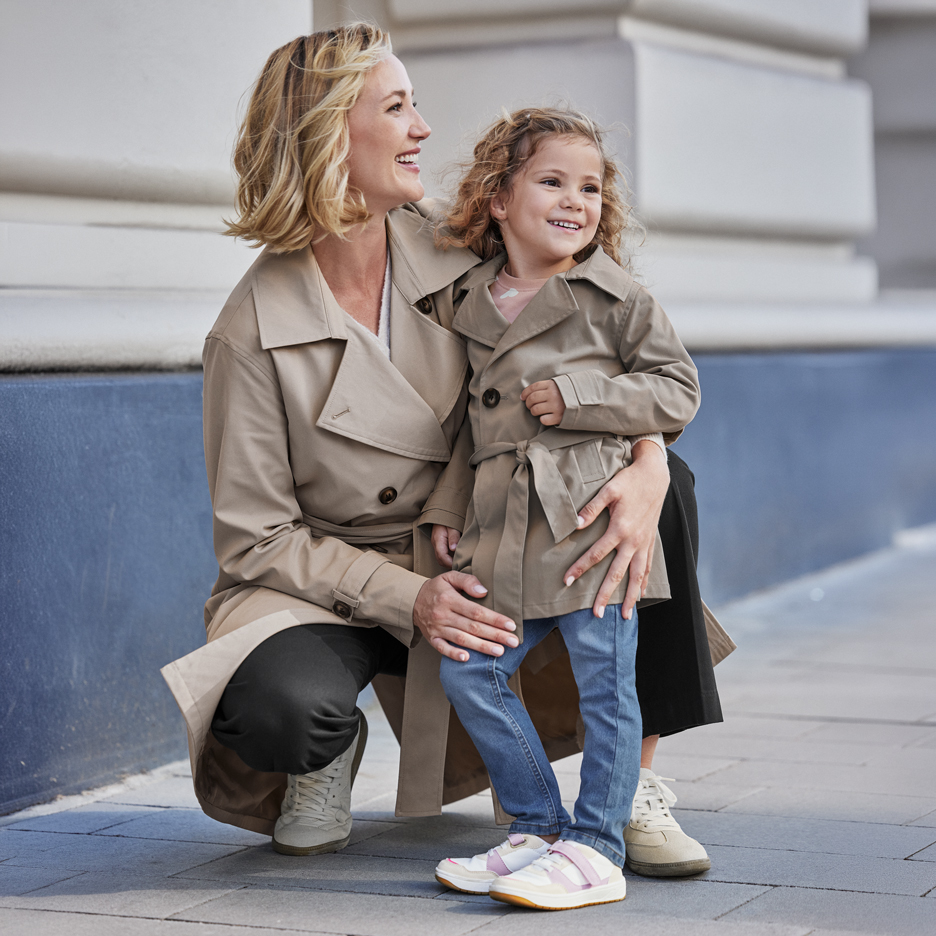Twee vrouwen in beige trenchcoats, een volwassene en een kind, lachend buiten.