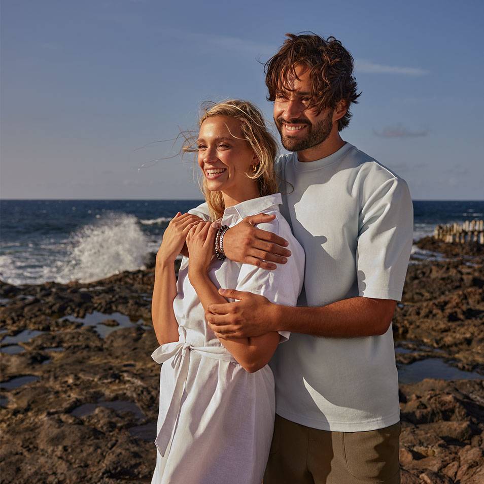 Man en vrouw in vrijetijdskleding aan de kust, man draagt een T-shirt, vrouw een jurk.
