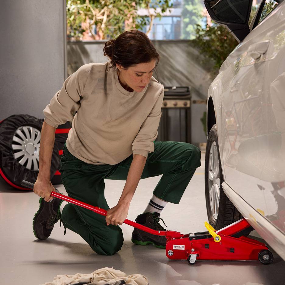 Vrouw gebruikt een rode hydraulische krik om een witte auto op te tillen, met een reservewiel in de buurt.