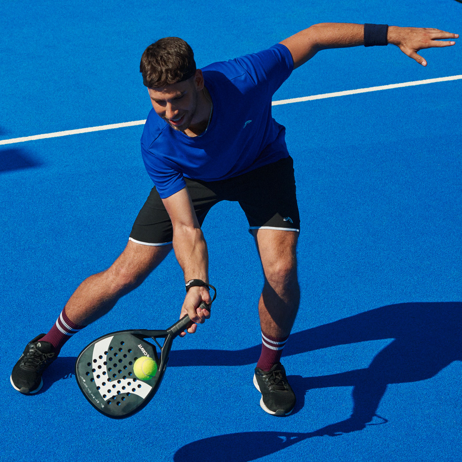 Man in blauw T-shirt en zwarte korte broek speelt padel met racket en bal.