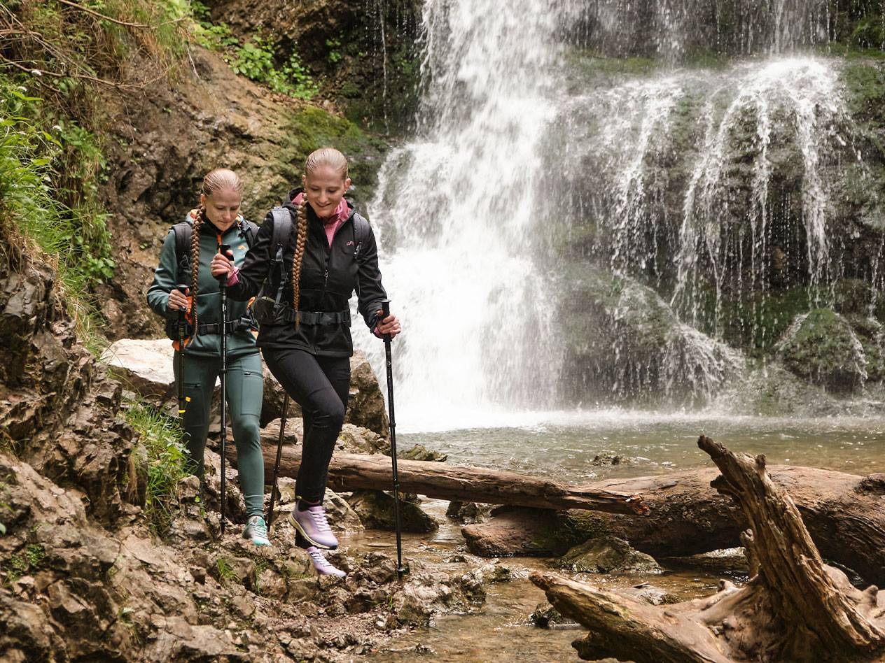 Deux femmes avec des bâtons de trekking marchent sur un sentier rocheux près d'une cascade.