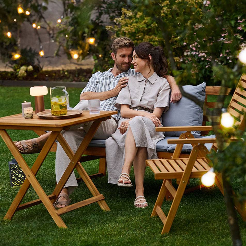 Een stel geniet van een drankje aan een houten tuintafel en bank in een tuin met sfeerverlichting.